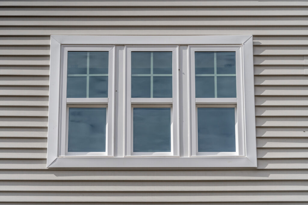 Triple hung window with fixed top sash and bottom sash that slides up, sash divided by white grilles a surrounded by white elegant frame horizontal white vinyl siding on a new construction residence